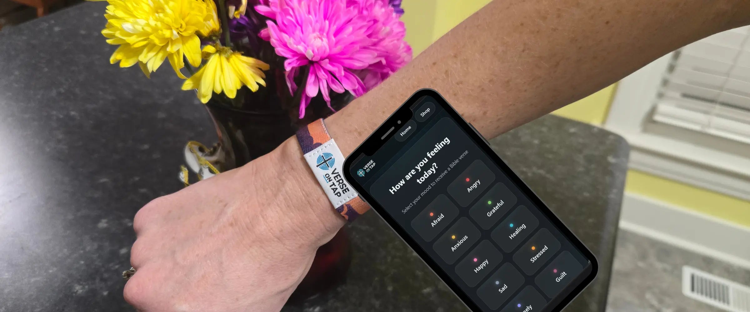 A close-up of a person's wrist wearing a white and orange smart bracelet with a smartphone attached or overlaid, showing a dark app screen, with yellow and pink flowers in the background on a dark countertop.